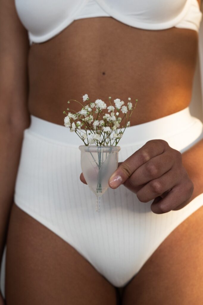 Quadril de uma mulher, que está de calcinha clara, com as mãos na frente do corpo segurando um coletor menstrual com uma flor branca dentro.